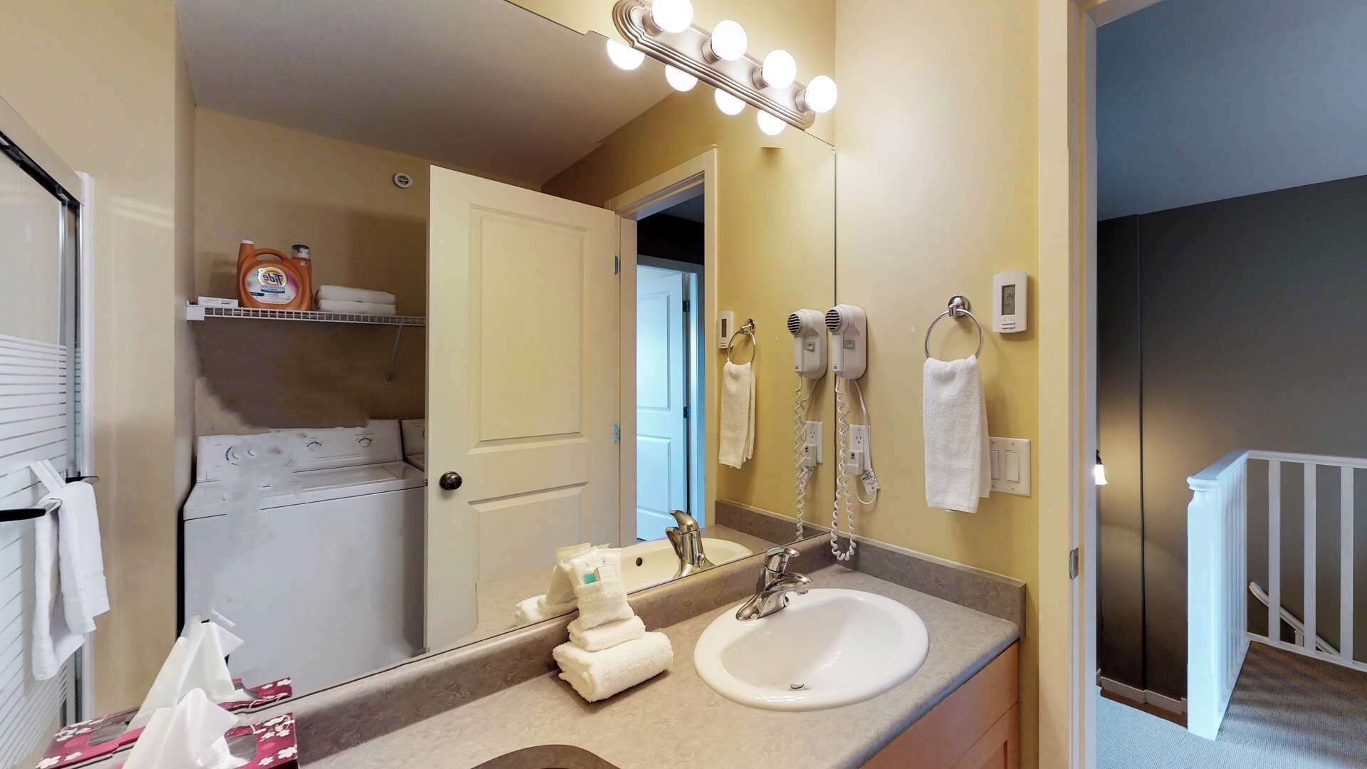 A hotel bathroom with a sink, mirror, and hairdryer. There is a laundry area with a washing machine and a shelf with toiletries.