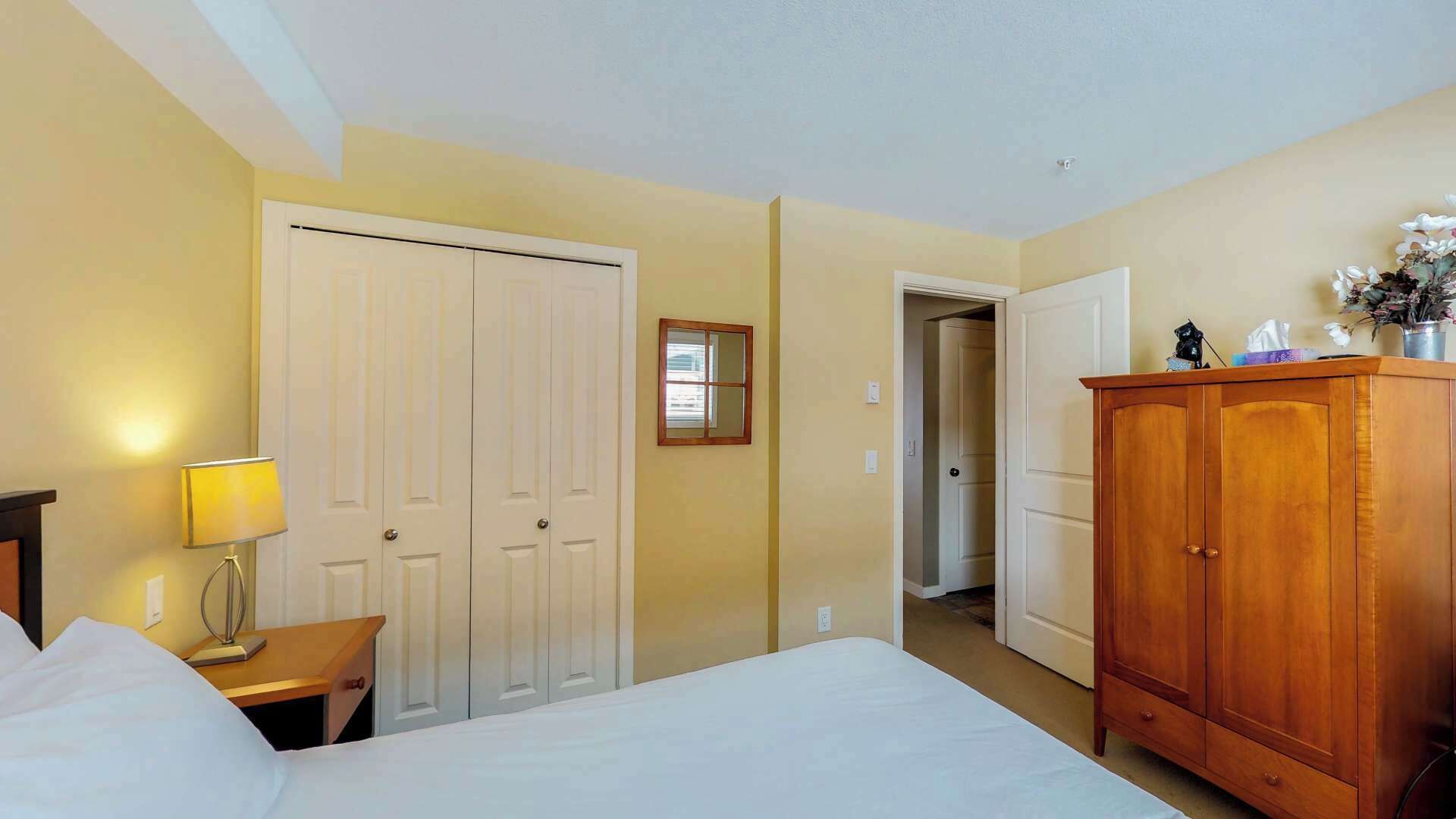 Une chambre d’hôtel avec un lit, une armoire en bois, une table de chevet avec une lampe, une porte et une petite fenêtre.