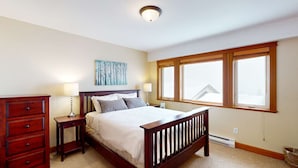 A bedroom with a wooden bed, two bedside tables, a dresser, and a large window with a view. - Okanagan - Tree Tops 10 (Big White Mountain)
