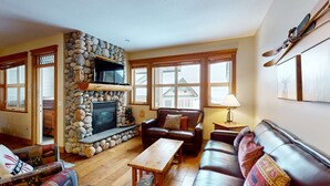 A cozy living room with a stone fireplace, leather sofas, a wooden coffee table, and large windows. - Okanagan - Tree Tops 10 (Big White Mountain)