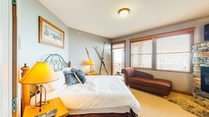 A bedroom with a bed, two bedside lamps, a chair, a fireplace, and a window with blinds. - Okanagan - Tree Tops 13 (Beaverdell)