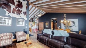 Ein geräumiges Wohnzimmer mit einer Ledersofa, einem gemusterten Sessel und einem Couchtisch aus Holz. Im Hintergrund ist eine Küche erkennbar, und die Decke weist freiliegende Holzbalken auf.