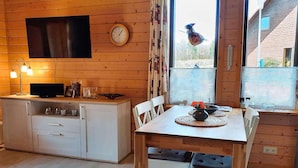 L’intérieur chaleureux d’un chalet, avec un revêtement mural en bois, une table à manger entourée de chaises, un coin cuisine équipé d’un téléviseur et une fenêtre donnant sur le paysage extérieur.