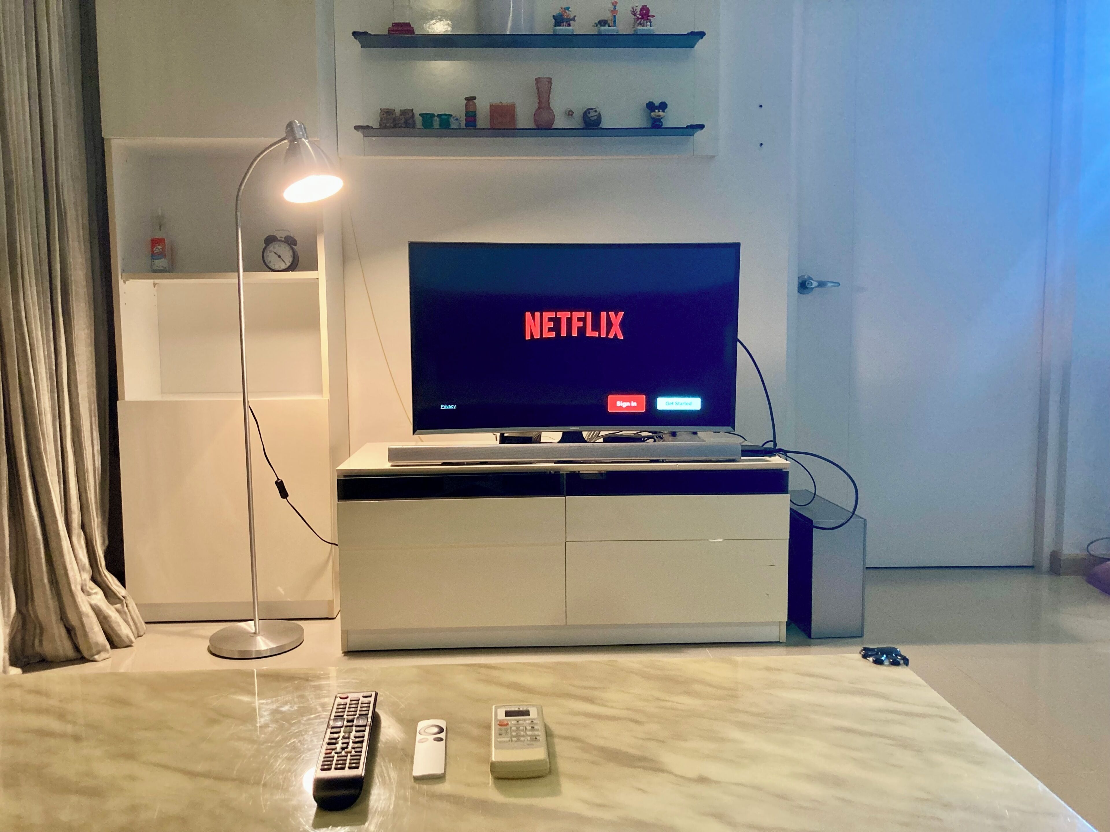 Un televisore con il logo Netflix, un telecomando, una mensola a parete con oggetti decorativi e una lampada da terra.