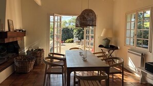 Een eetruimte met een houten tafel en stoelen, een hanglamp, een open haard en uitzicht op de tuin door openslaande deuren.