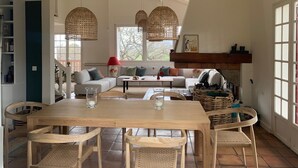 Een ruime woonkamer met een houten eettafel, rieten stoelen, een bank en een open haard.