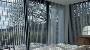Une chambre avec un grand lit, un fauteuil en osier, une table de chevet en bois agrémentée d’un objet décoratif, et une vue dégagée grâce à de grandes fenêtres du sol au plafond.