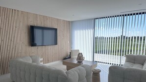Un salon moderne doté d’un grand téléviseur à écran plat fixé à un mur en bois, d’un canapé, d’une table basse et offrant une vue sur un paysage verdoyant grâce à de grandes fenêtres du sol au plafond.