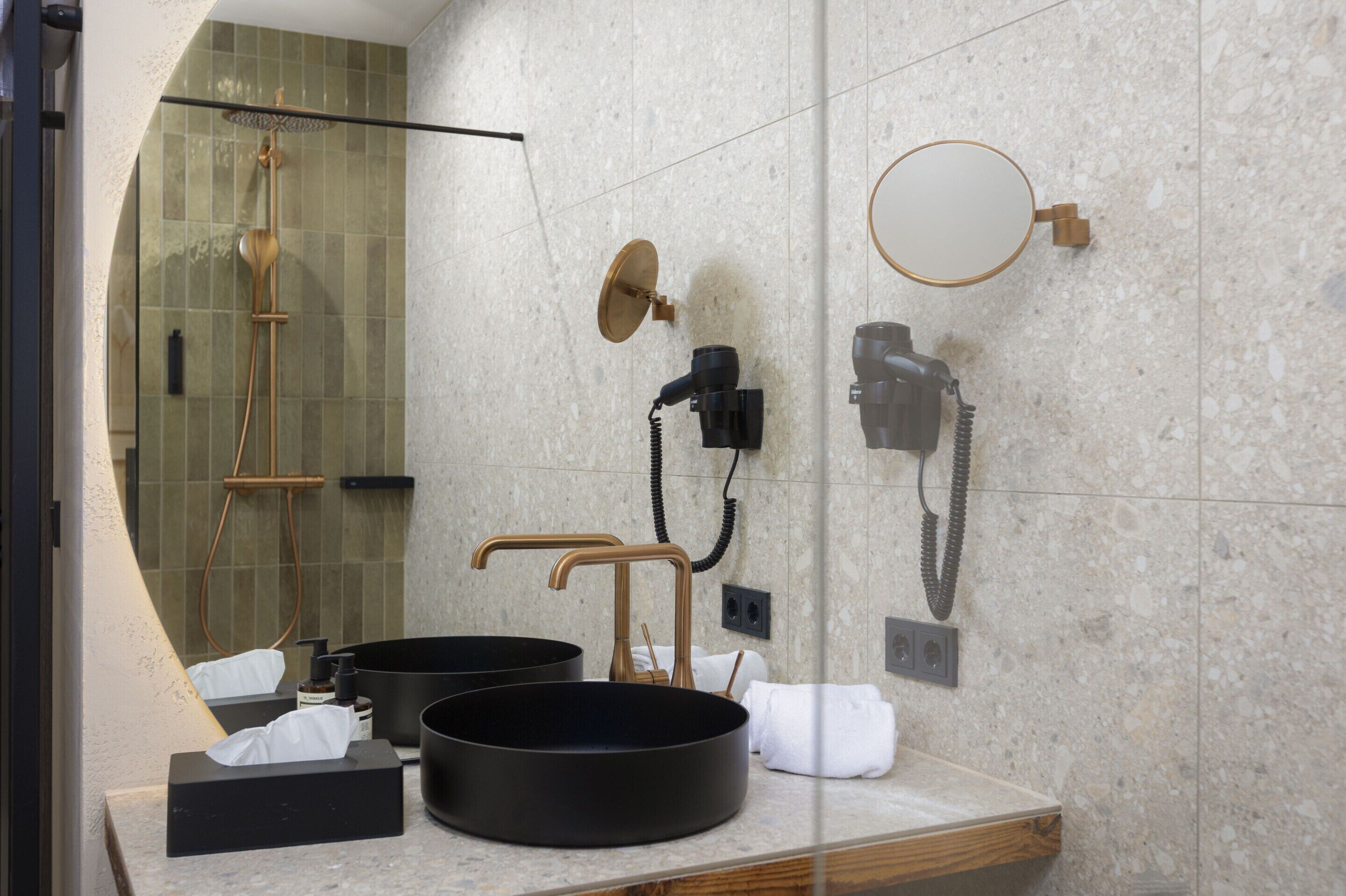 Un baño moderno con ducha acristalada, dos lavamanos, un espejo redondo y un secador de pelo montado en la pared.