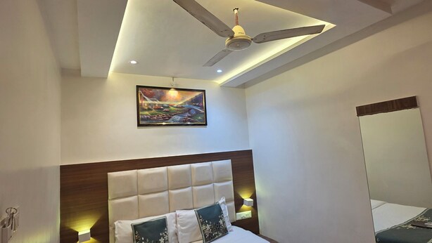 A modern hotel room with a large bed, a ceiling fan, a nightstand, a wall-mounted lamp, and a painting above the bed. - Hotel Wellingten (Mumbai)