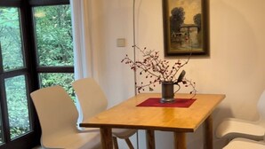 Un comedor con una mesa de madera, dos sillas blancas, un mantel individual rojo, un florero metálico con ramas, una lámpara de pie y un cuadro enmarcado en la pared.