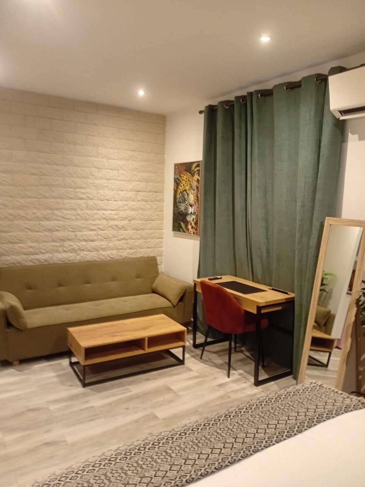 Una habitación amoblada con un sofá, una mesita de madera, un escritorio con una silla, un espejo y un tapete estampado.