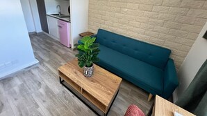 Un espacio habitable pequeño con kitchenette, un sofá azul, una mesa de centro de madera con una planta y una pared con diseño de ladrillo.