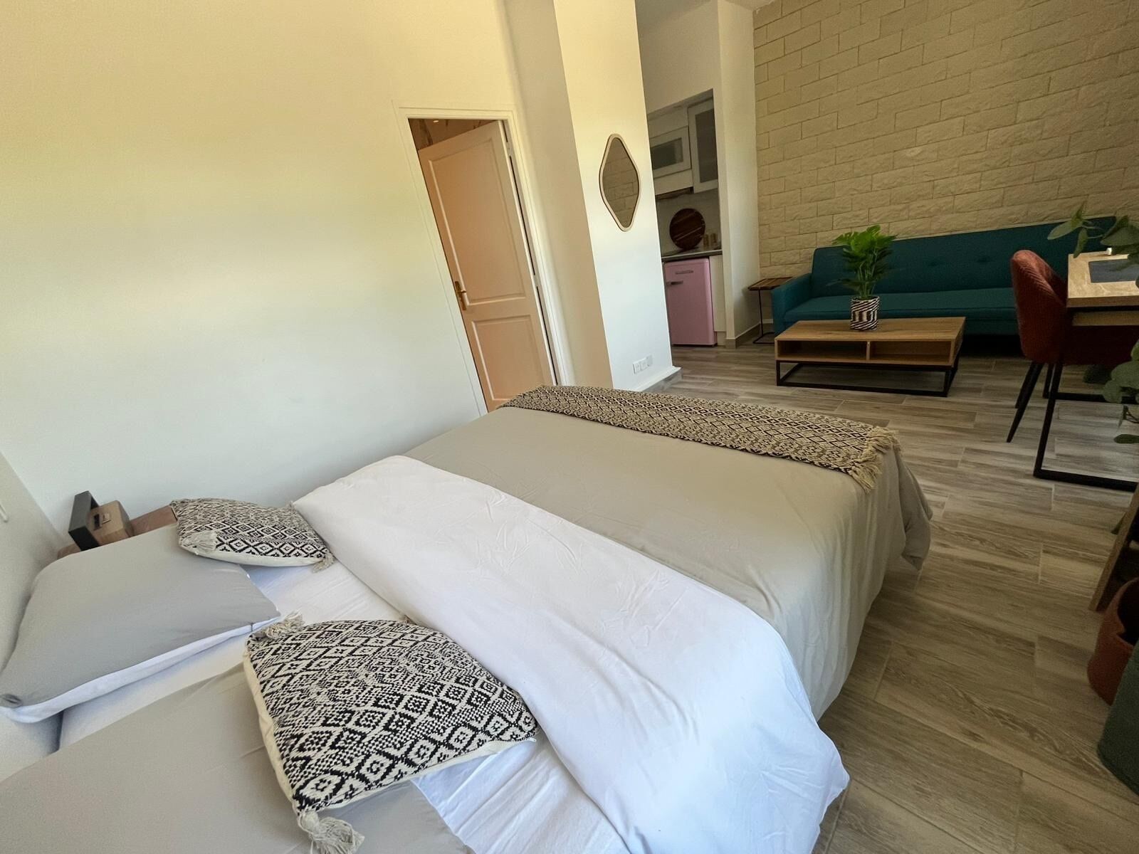 A bedroom with two beds, a wooden floor, a green couch, a wooden table, and a kitchen area in the background.