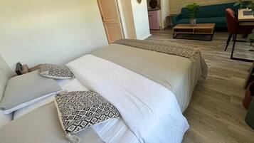 A bedroom with two beds, a wooden floor, a green couch, a wooden table, and a kitchen area in the background.