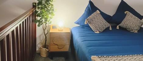 Un dormitorio con techo inclinado, vigas de madera, cama con ropa de cama azul, almohadas estampadas, una lámpara de noche y una planta en maceta.