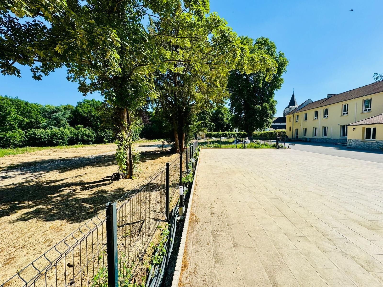 A paved courtyard with a fence, trees, and a building with a tiled roof.