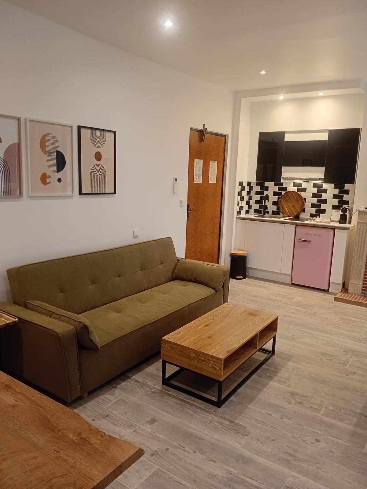 Ein Wohnzimmer mit Sofa und Couchtisch, eine Küche mit gefliestem Fliesenspiegel und einem pinken Kühlschrank sowie drei gerahmte Kunstwerke an der Wand.