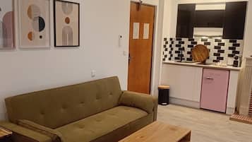 Ein Wohnzimmer mit Sofa und Couchtisch, eine Küche mit gefliestem Fliesenspiegel und einem pinken Kühlschrank sowie drei gerahmte Kunstwerke an der Wand.