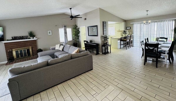 A spacious living room with a grey sectional sofa, a fireplace, a dining area with a table and chairs, and a kitchen in the background.