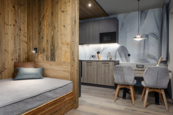 Ein modernes Zimmer mit einer Wand in Holzoptik, einem Bett mit grauem Topper, einer kleinen Küchenzeile mit Mikrowelle und zwei grauen Stühlen.