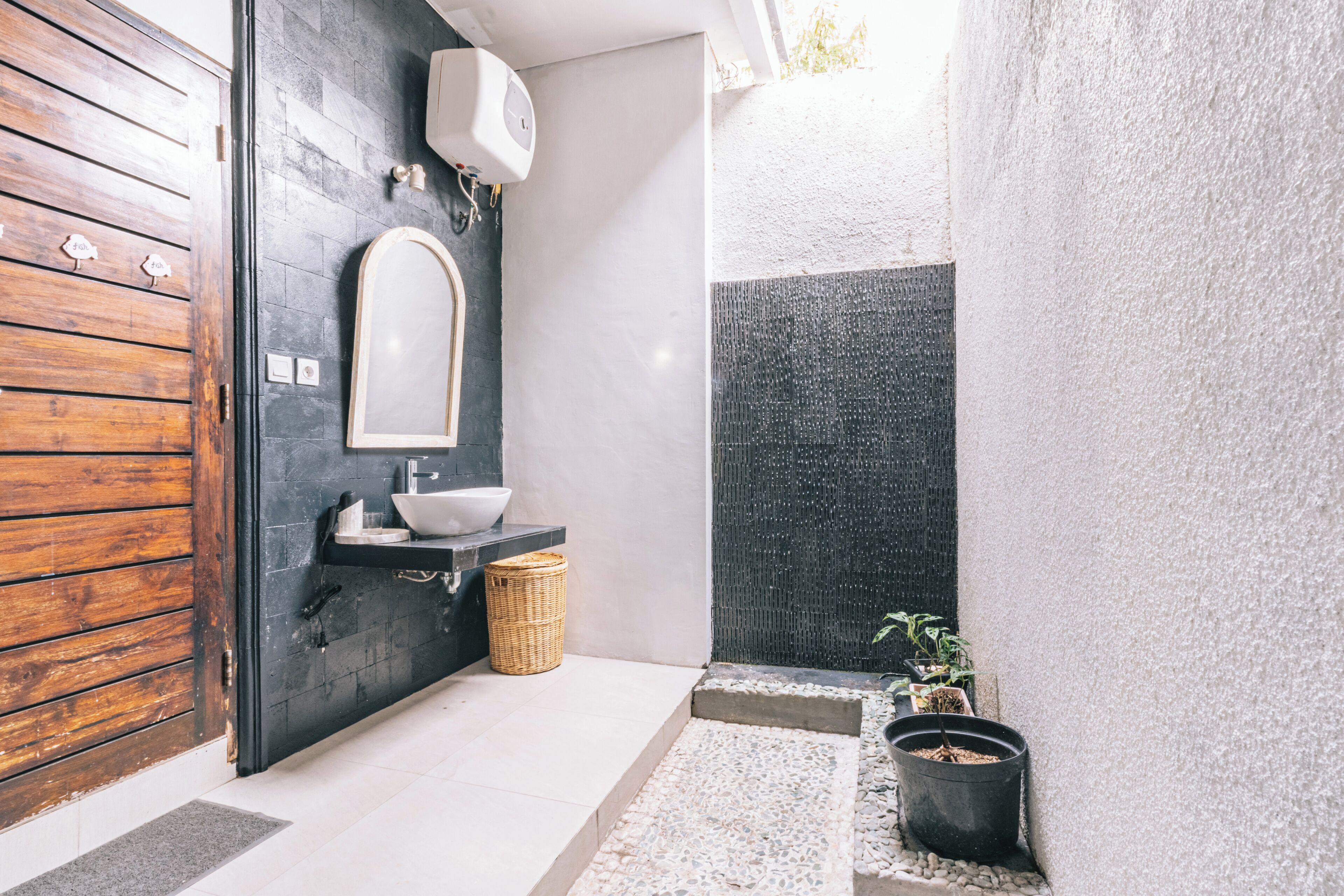 Un baño moderno con puerta de madera, un lavamanos blanco, un espejo, una pared negra con textura y una pequeña planta en maceta.