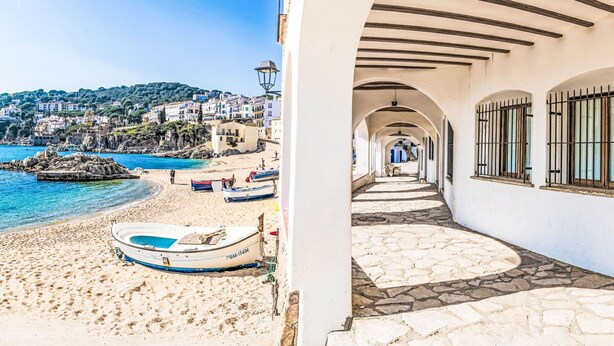 Um edifício costeiro com um caminho de pedra que leva a uma praia com barcos e um mar azul cristalino.