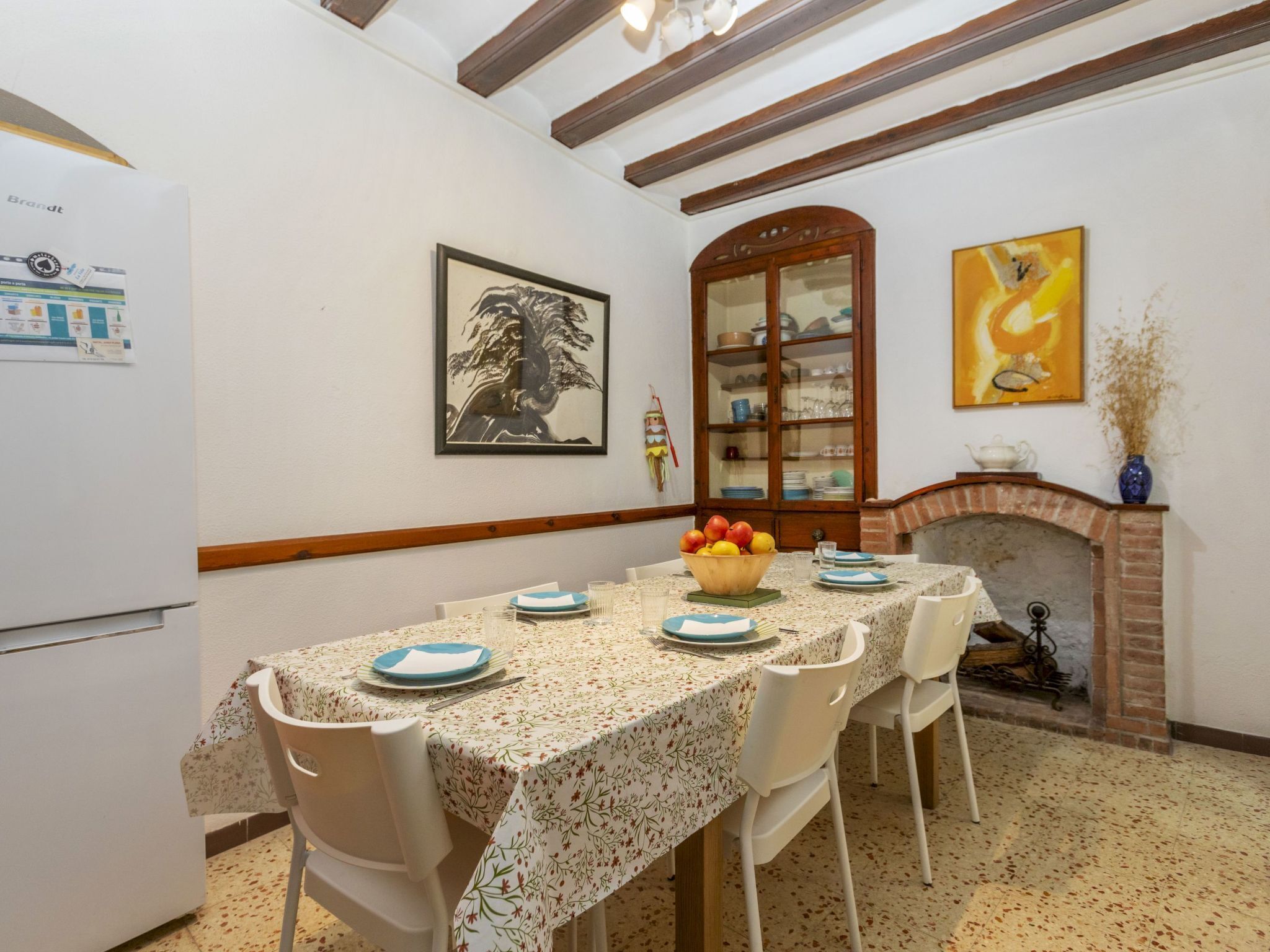 Un coin repas avec une table dressée pour quatre personnes, entourée de chaises blanches. Un buffet en bois, doté de portes en verre, est adossé au mur, et on aperçoit une cheminée au fond.