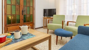 Uma sala de estar com uma mesa de madeira, duas cadeiras, uma televisão e uma janela grande com cortinas.