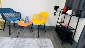 Une pièce avec deux chaises, une petite table, un tapis blanc et moelleux, une étagère noire sur laquelle sont posés divers objets, et une prise électrique au mur.