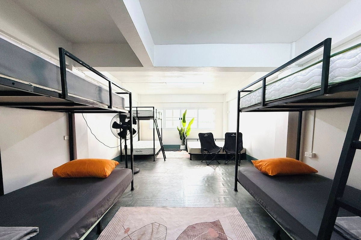 A dormitory room with bunk beds, a desk, a chair, a fan, and a window.