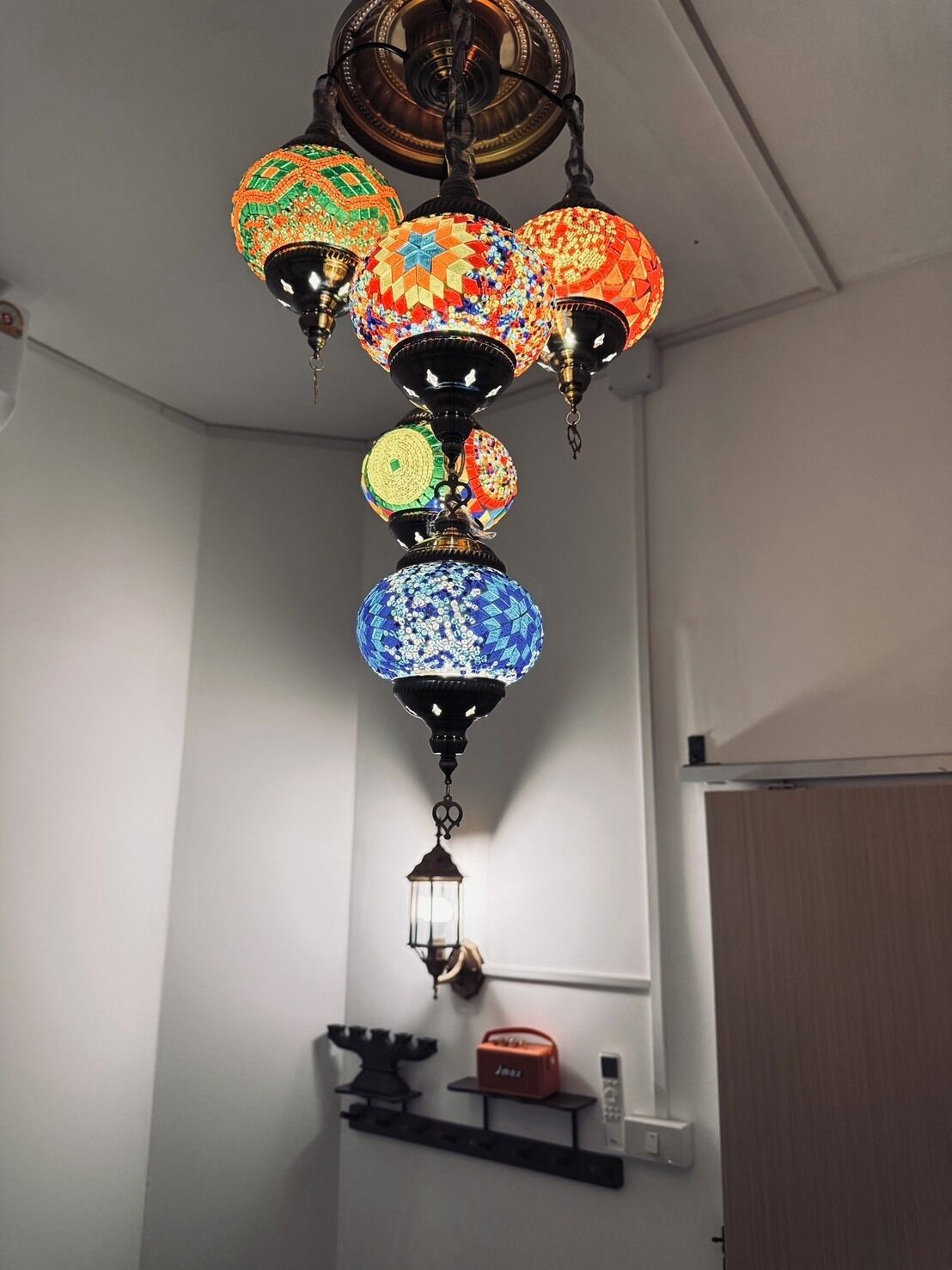 A multi-tiered hanging lamp with a traditional design, a wall-mounted shelf with a small speaker and a remote control, and a door in the background.