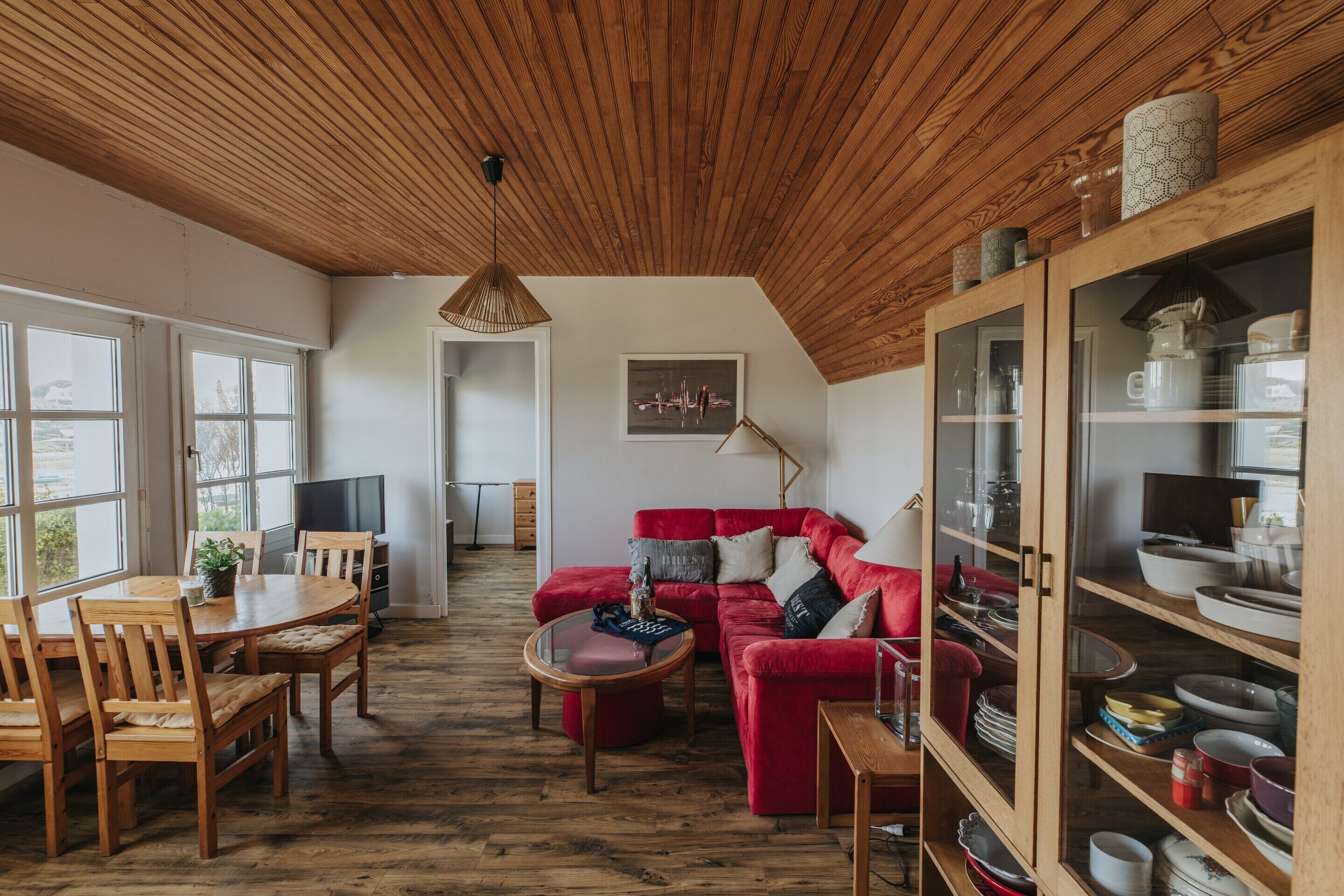 Una sala de estar acogedora con un sofá rojo, una mesa de comedor de madera y sillas. Hay un televisor, una lámpara y un aparador con vajilla.