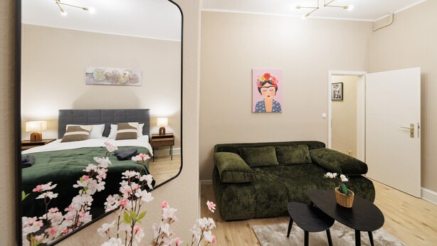 Quarto de hotel moderno com um espelho grande, um sofá verde, uma mesinha e uma cama com colcha verde.