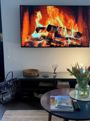 Ett vardagsrum med en platt-TV som visar en bild av en öppen spis, ett soffbord med böcker och en fjärrkontroll, en liten vas med blommor och en korg av rotting.