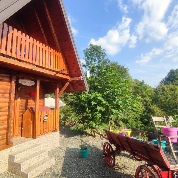 A wooden cabin with a porch, a wooden cart, and a sign reading "Bajat Octof".