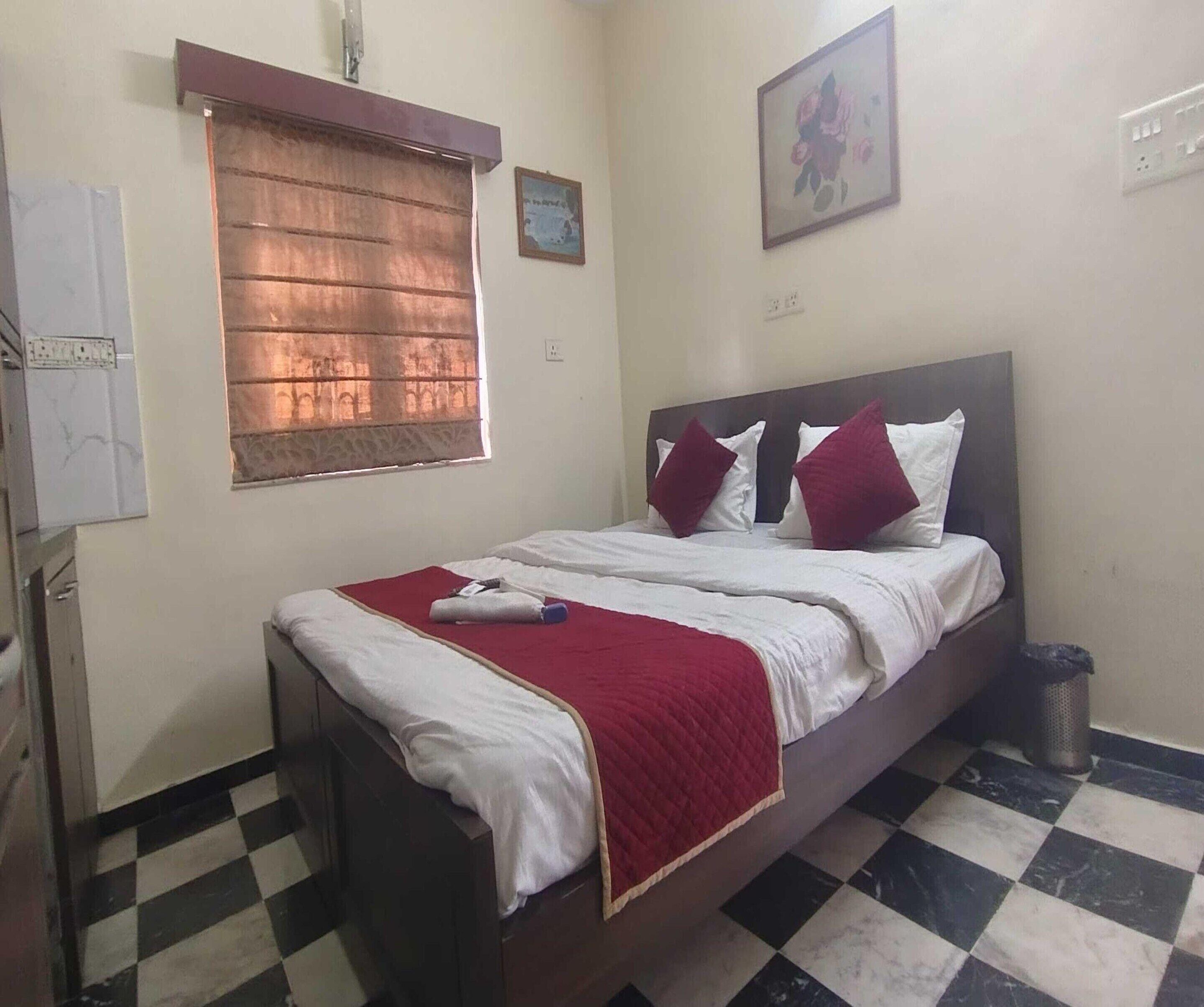 A single bed with a wooden headboard, a red throw blanket, and two red pillows. There is a window with a brown curtain, a wall-mounted picture, and a trash bin beside the bed.