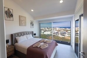 A modern bedroom with a large bed, a view of a cityscape through a floor-to-ceiling window, and a balcony with a glass door. - Protaras Holiday Villa Ds112 (Paralimni)