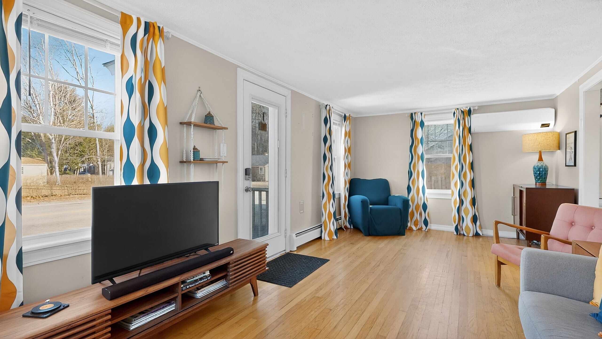 Una sala de estar con televisor, un sillón azul y una silla rosa. Se observan cortinas estampadas y una lámpara sobre una mesita.