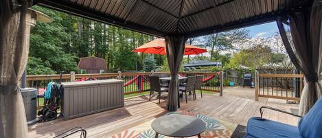 Una terraza techada al aire libre con zona de estar, una parrilla y vistas al bosque circundante.
