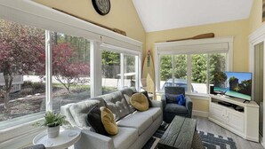 Una sala de estar con sofá, televisor y ventanas con vistas a un jardín.