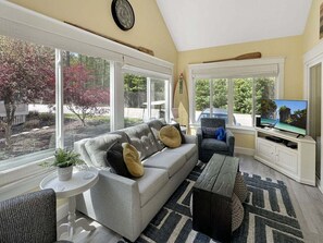 Una sala de estar con sofá, televisor y ventanas con vistas a un jardín.