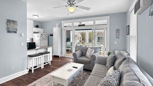 Ein modernes Wohnzimmer mit grauem Sofa, weißem Couchtisch und einer Küchenzeile mit weißen Schränken und einem Kühlschrank.