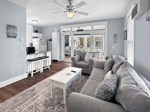 A modern living room with a gray sofa, white coffee table, and a kitchen area with white cabinets and a refrigerator. - Seaglass Village 172 - Seaside Serenity Sanctuary (Wells)