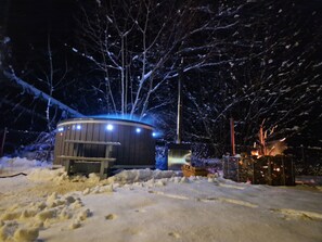 Ein verschneiter Außenbereich mit einer Holzhütte, die von Lichtern erhellt wird, umgeben von schneebedeckten Bäumen.