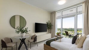 A modern hotel room with a large bed, a flat-screen TV, a round mirror, a small table with chairs, and a potted plant. - Refined Studio w/ Top Facilities in Damac Hills (Dubai)