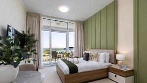 A modern hotel room with a large bed, a bedside table with a lamp, a small table with chairs, and a potted plant. - Refined Studio w/ Top Facilities in Damac Hills (Dubai)