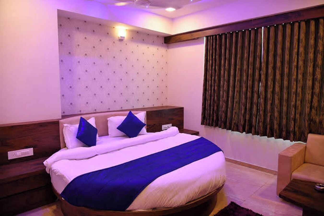A hotel room with a large bed, a headboard with a tufted design, a wooden nightstand, a bedside table, a chair, and a window with curtains.