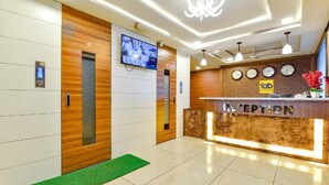 A hotel reception area with a reception desk, a television mounted on the wall, and a clock display. - Hotel PVR (Ahmedabad)