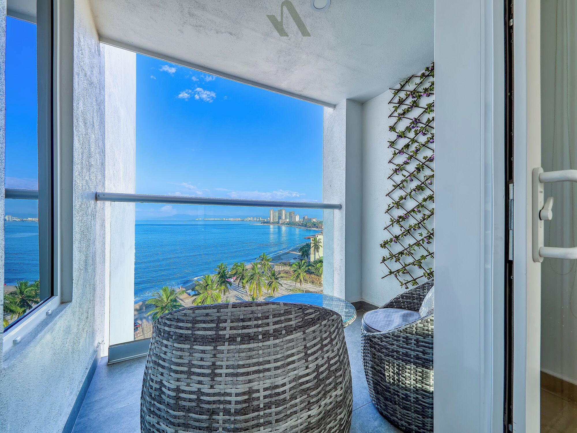 A balcony with wicker furniture, a view of the sea, and a cityscape in the background.